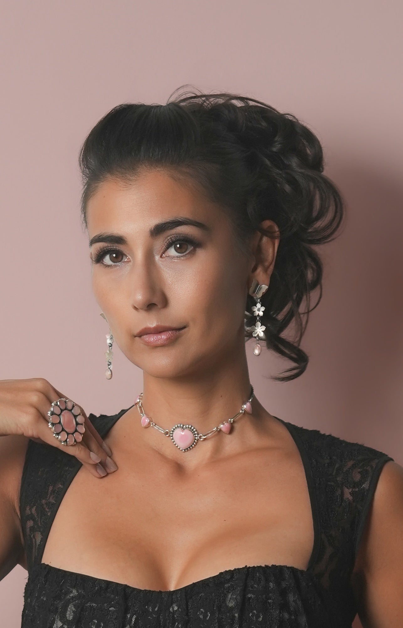 A woman with dark hair styled in an updo models a pink and silver heart-themed jewelry set, including a necklace, matching drop earrings, and a ring, against a solid dusty pink background.