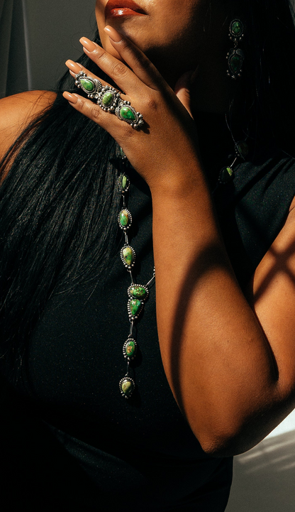 Woman wearing a black dress with green jewelry against a gray wall.
